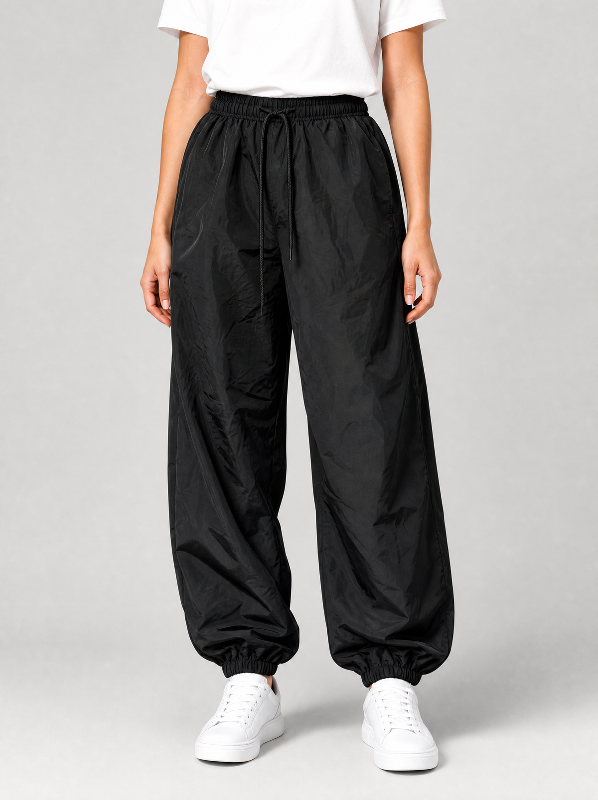Woman wearing black parachute pants with cuffed hem, front view on a light grey studio background
