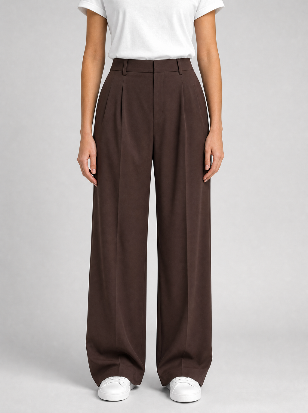 Woman wearing brown pleated high waist wide leg trousers, front view on a light grey studio background