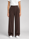 Woman wearing brown pleated high waist wide leg trousers, front view on a light grey studio background