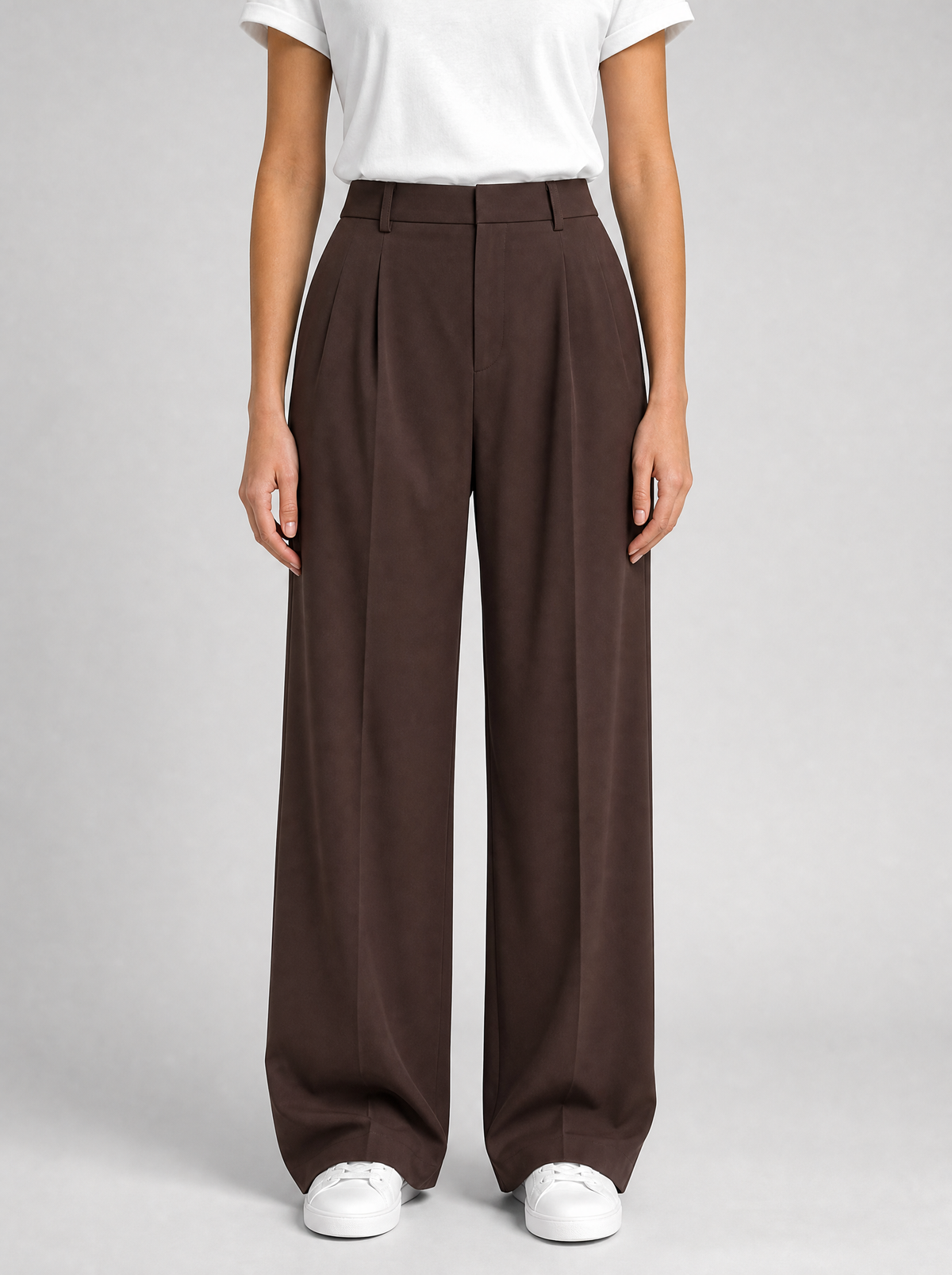 Woman wearing brown pleated high waist wide leg trousers, front view on a light grey studio background
