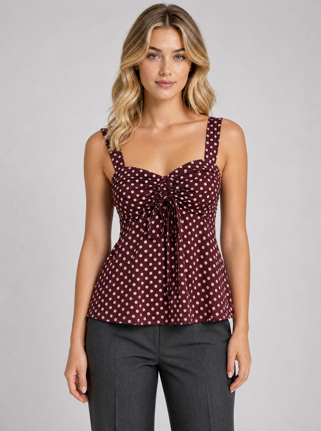 Woman wearing a burgundy polka dot ruched cami blouse, front view on a light grey studio background